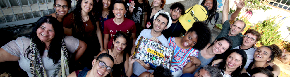 Diversas e diversos jovens reunidos em torno de um jovem que segura uma maleta com a palavra "Juventudes" nela.
