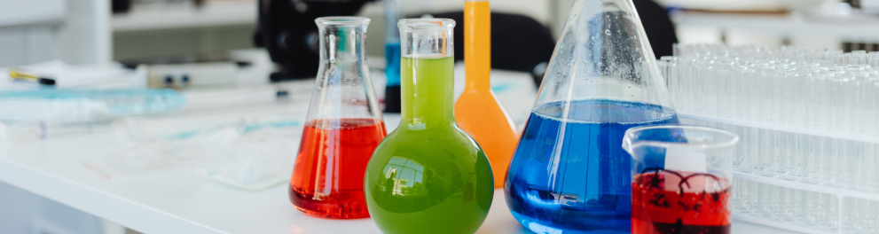 Diversos frascos com líquidos coloridos dispostos sobre uma bancada branca, em um laboratório.