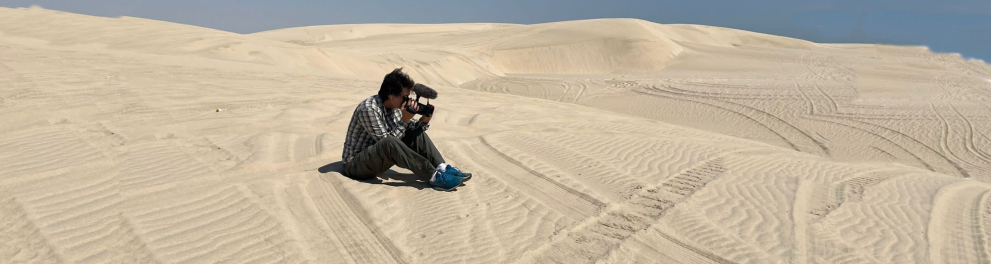 Homem está sentado sobre a areia de dunas, usando uma câmera. Ao fundo, o céu azul.