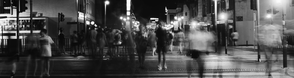 Imagem em preto e branco, com pessoas e vultos atravessando uma rua à noite.