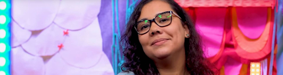 Jovem morena usando óculos e uma camisa azul com uma blusa preta por baixo com um cenário colorido ao fundo