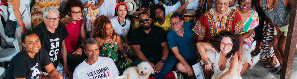 Diversas pessoas negras estão reunidas, algumas sentadas, outras de pé, para celebração da estreia do documentário "Mimbó".