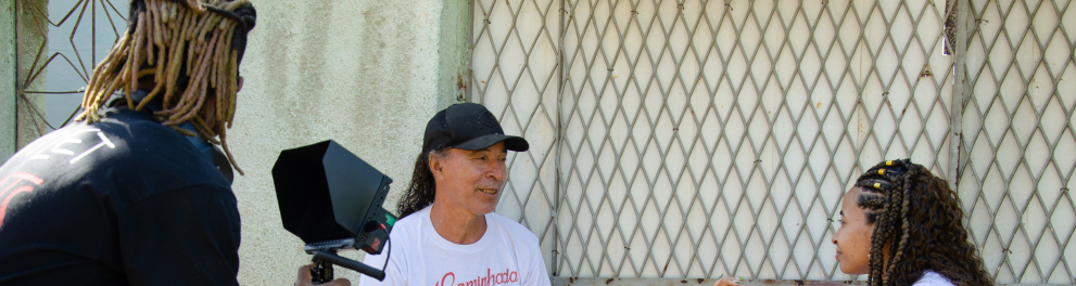 Homem negro segura uma câmera e grava duas pessoas sentadas