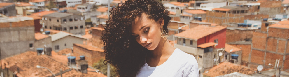 Uma mulher negra está com seu cabelo (longo e cacheado), olhando para baixo. Ao fundo, casas.