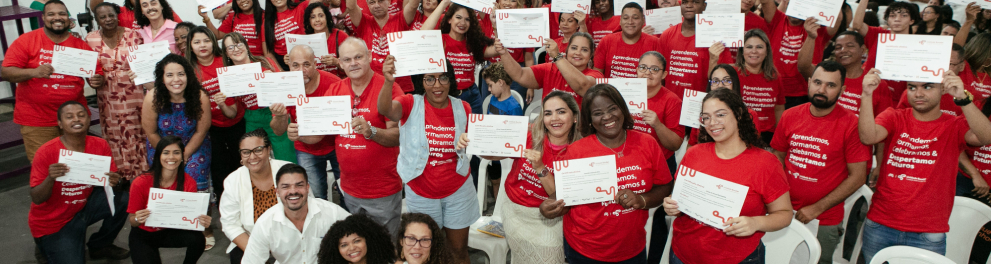 Diversas pessoas usando camisas vermelhas seguram, sorrindo, seus certificados de conclusão dos estudos no ensino fundamental e no ensino médio.