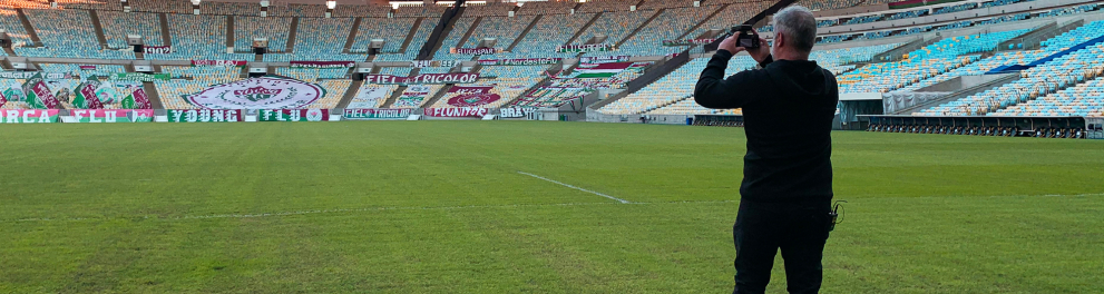 Homem está de pé no gramado de um estádio de futebol. Ele parece fotografar (ou filmar) a arquibancada.