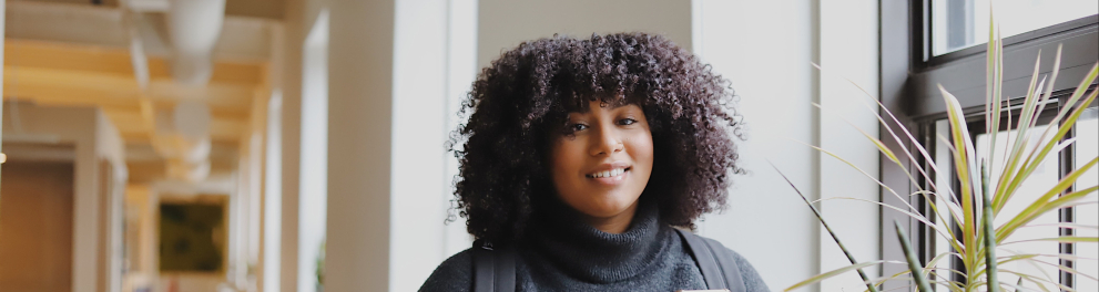 Uma jovem negra próxima a uma janela mexendo no celular e sorrindo para uma foto