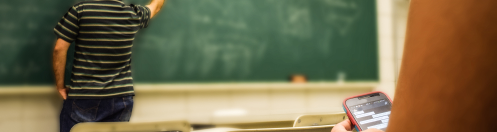 Um professor está escrevendo no quadro enquanto uma estudante segura um celular.