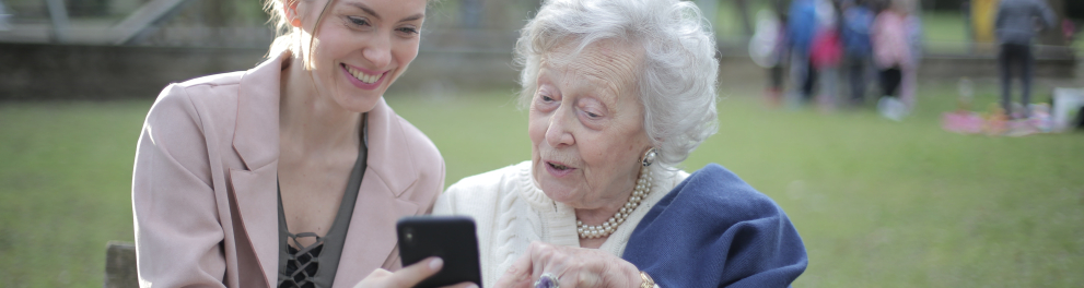 Duas mulheres estão em um banco de praça. A mais nova mostra a tela do celular para a mais velha. Ambas sorriem.