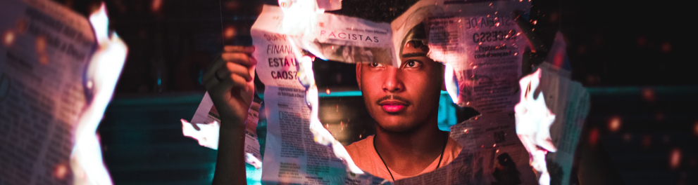 Homem segura um jornal que pega fogo e tem sua parte central já totalmente queimada e aberta.