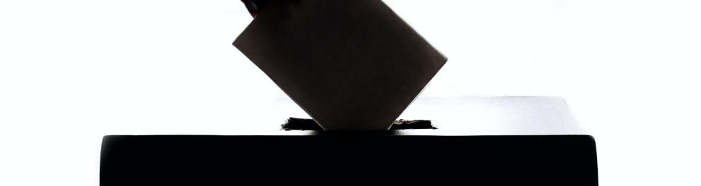 A imagem mostra uma mão colocando um papel em uma urna