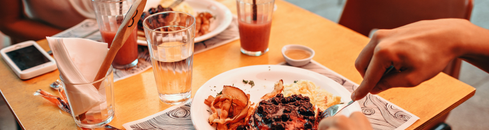 Duas pessoas estão diante de seus pratos de comida sobre a mesa de um restaurante a céu aberto. Seus rostos não estão enquadrados na foto.