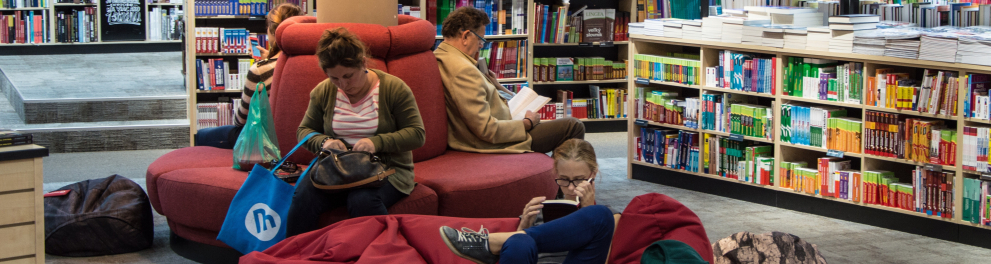 Pessoas estão sentadas em sofás, dentro de uma biblioteca, lendo.