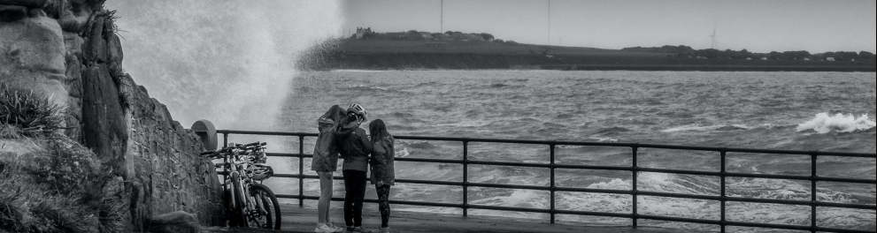 Imagem em preto e branco. Três jovens assistem ao bater das ondas. Ao lado dos três, suas bicicletas.