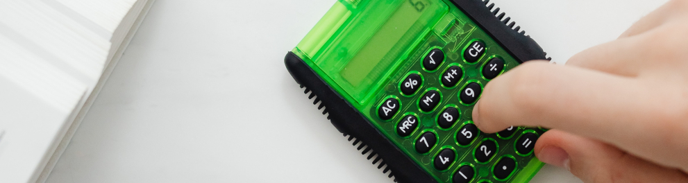 Imagem mostra um Dedo sobre calculadora em cima da mesa e caderno ao lado