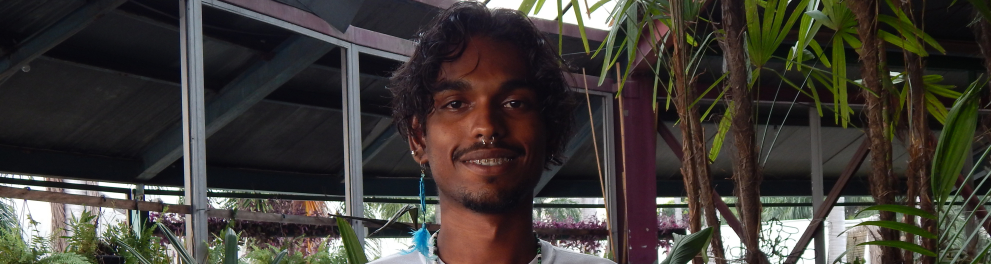 Jovem sorri. Ele está de pé, usando uma camisa branca. Ao fundo, plantas.