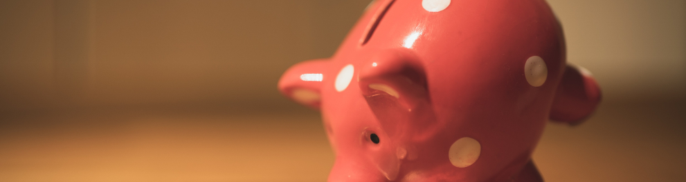 Imagem de um cofrinho na forma de um porquinho cor de rosa. O porquinho está em cima de uma mesa com algumas moedas em sua frente