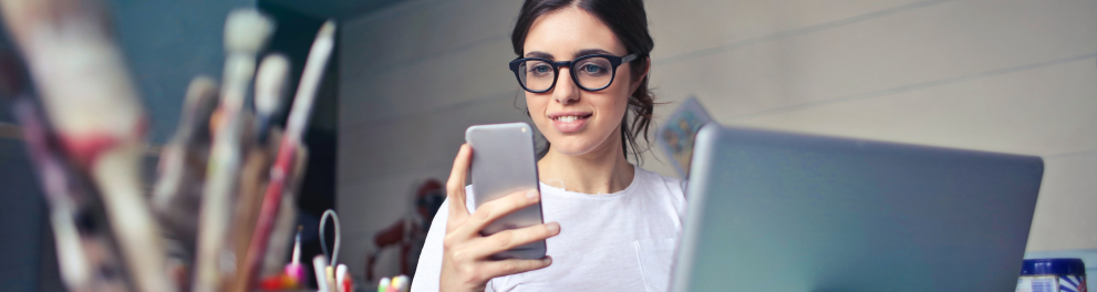 Jovem mulher de óculos, sentada em frente a um notebook, porém mexendo e olhando sorridente para o celular em sua mão direita