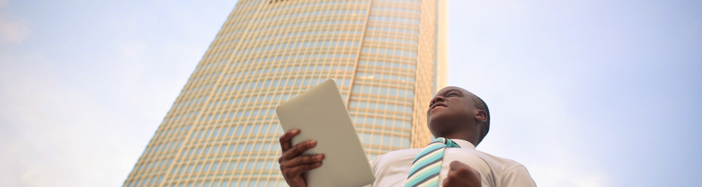 Um jovem negro de camisa social, gravata e calça social está à frente de um edifício de cerca de uns 150 andares. O jovem está segurando um tablet com a mão direita e comemorando algo com a mão esquerda e uma expressão de felicidade no rosto