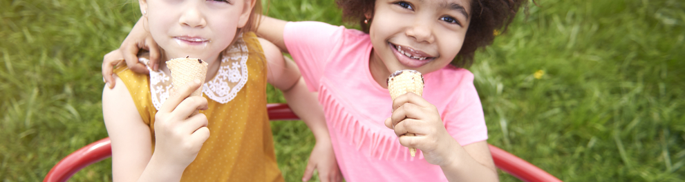 Duas meninas de cerca de 3 anos de idade - uma negra e uma branca - estão rindo para a foto, segurando um sorvete cada uma e abraçadas ao mesmo tempo