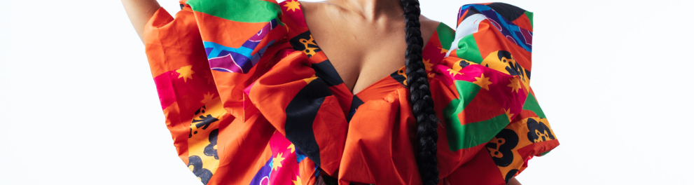 A artista negra Héloa trajando um vestido coloridíssimo, ostentando uma longa trança e com os braços abertos à frente um fundo totalmente branco