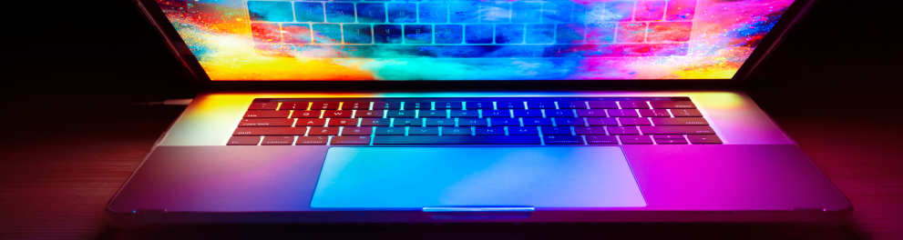 Imagem de um notebook semi aberto em cima de uma mesa. O notebook está ligado e com uma tela bem colorida que está iluminando a mesa