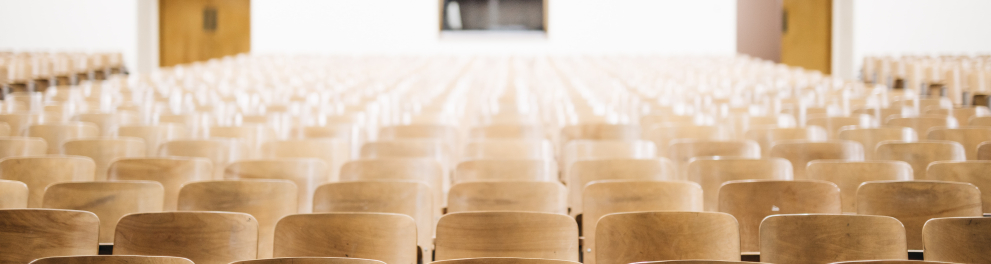 Foto de um auditório com as cadeiras vazias na cor de madeira. Ao fundo, duas portas de saída e uma janela de uma sala de controle de comunicação do auditório