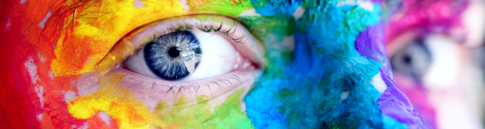 Close nos olhos azúis de uma pessoa que está olhando para a câmera e tem o rosto pintado nas cores da bandeira de arco-íris
