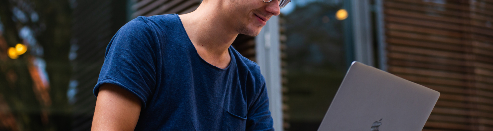 Um rapaz loiro, de óculos, usando calça jeans e camisa azul está sentado com um notebook aberto às mãos e olhando para o mesmo