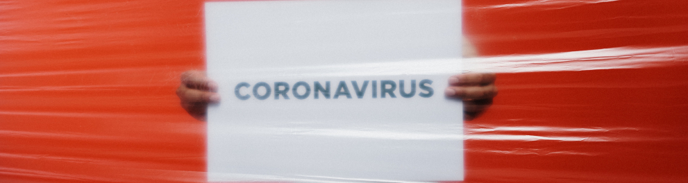 Um homem negro usando máscara de proteção contra a covid-19 está segurando uma folha de papel com a palavra "coronavirus" á mostra para a câmera. Entre o homem e a câmera há um muro de plástico