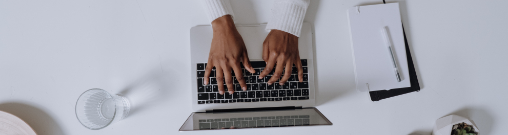 Um notebook aberto em cima de mesa branca. Duas mãos estão teclando as teclas do notebook e a imagem foi feita de um take aéreo