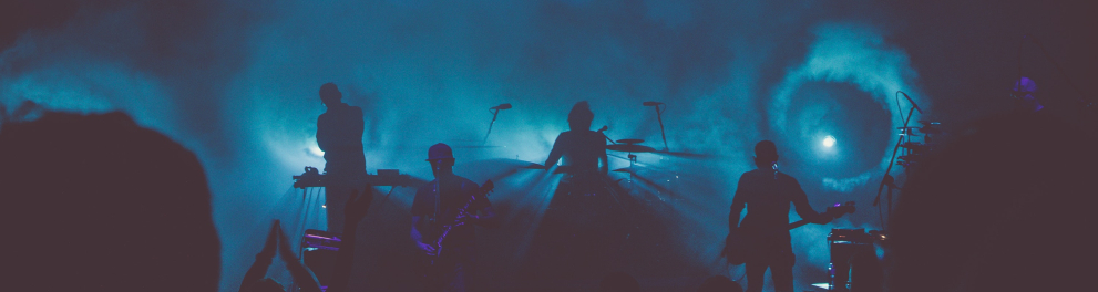 Imagem escura, mostrando apenas as silhuetas de quatro músicos em um palco com iluminação azul