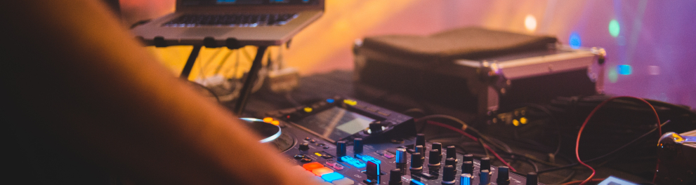 Mão de um DJ mexendo em aparelhagem de música eletrônica. Ao fundo, luzes coloridas mostram que é um ambiente de boate