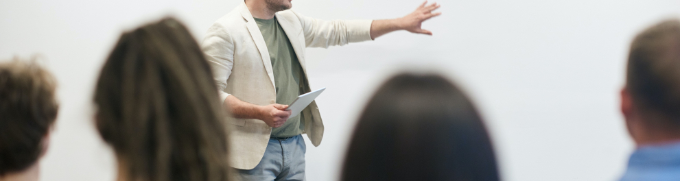 Professor de perfil apontando para o quadro em sala de aula enquanto é observado quatro alunos, que aparecem de costas na imagem