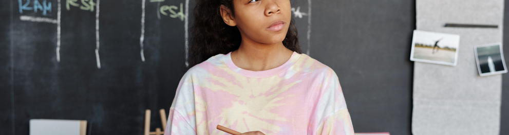 Menina negra com uma camisa cor de rosa segurando um caderno com a mão esquerda e pensativa no que vai escrever, segurando um lápis na mão direita. Ao fundo, um quadro negro de sala de aula