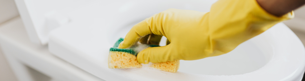 A mão de uma pessoa está com luva amarela de borracha esfregando a borda de um vaso sanitário branco com uma esponja