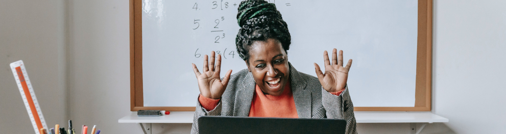 Professora negra sorridente com as mãos levantadas de frente a um notebook. Atrás dela está o quadro de sala de aula