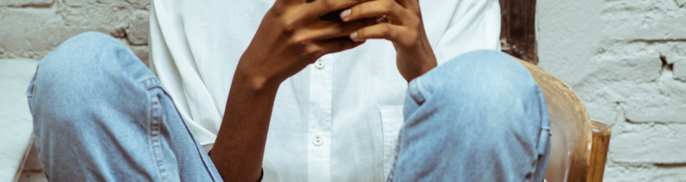 Uma jovem mulher negra com a cabeça raspada, usando óculos, calça jeans e blusa social branca está sentada em uma cadeira com as pernas também sobre a cadeira e mexeno num celular de forma séria