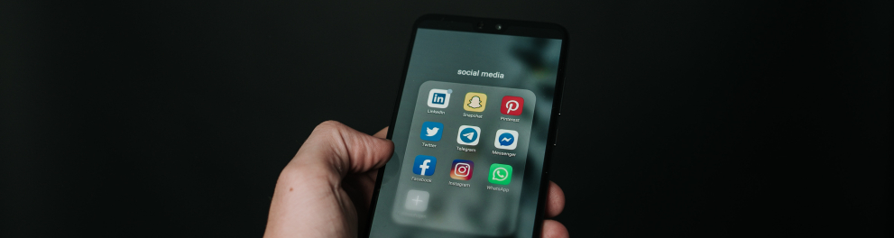 Uma mão masculina segurando um smartphone com a tela mostrando o ícone de nove apps de redes sociais. Fundo todo preto da imagem