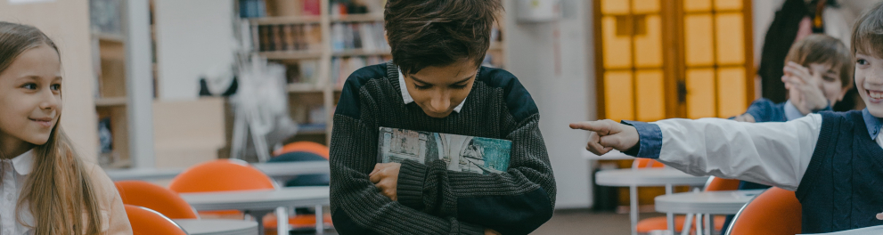Um menino em pé em sala de aula, cabisbaixo e abraçado aos livros enquanto outros alunos apontam os dedos e olham para ele rindo e debochando