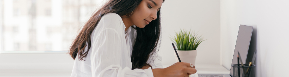 Uma adolescente mulata de cabelos lisos está de perfil sentada à uma mesa branca com o notebook aberto à sua frente e um caderno em que está escrevendo algo