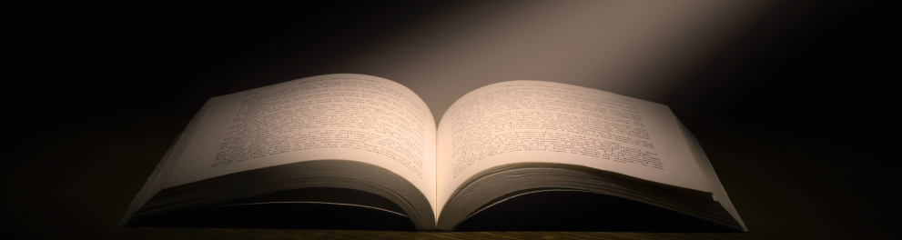 Imagem de um livro aberto em um fundo totalmente negro, porém com um raio de luz vindo do canto superior direito da imagem e batendo no centro do livro