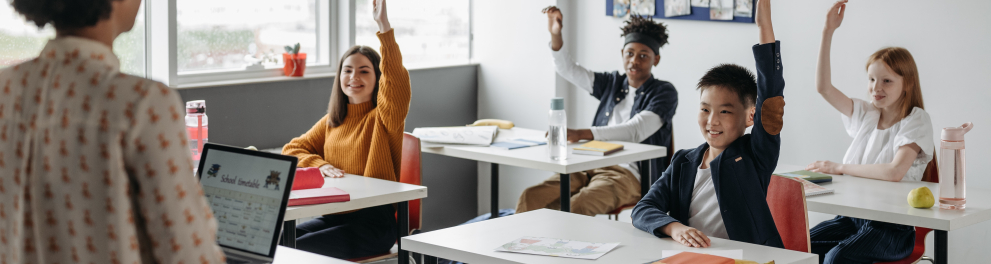 Sala de aula composta por quatro crianças levantando uma de suas respectivas mãos para fazer uma pergunta à professora, que está de frente para eles, em sua mesa de trabalho
