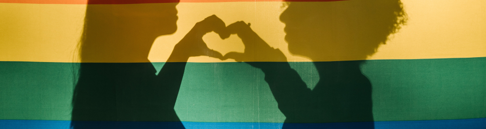 Sombras de duas mulheres fazendo um coração com as mãos situadas em uma bandeira de arco-íris LGBTQIA+