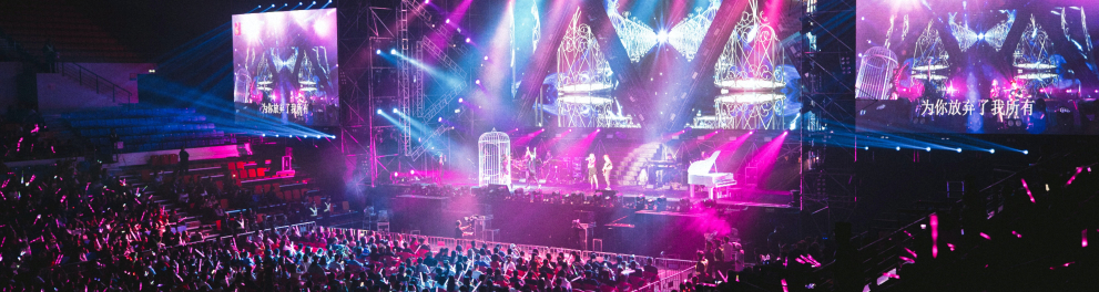 Imagem aberta de um show musical mostrando o palco bem colorido de azul e rosa e a multidão na plateia
