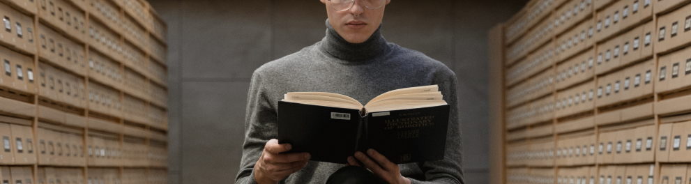 Homem sentado em uma cadeira de pernas cruzadas e segurando um livro, cujo o qual está lendo. O home usa óculos, está de calça preta, sapatos pretos e uma camisa cinza de gola rolê. Ao fundo, a imagem de santos