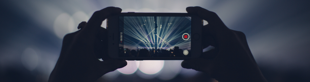 Celular sendo segurado na posição horizontal pelas duas mãos de uma pessoa, que está filmando um show musical