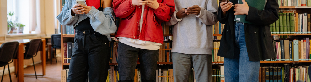Quatro estudantes em pé e um ao lado do outro, mexendo em seus respectivos celulares dentro de uma biblioteca