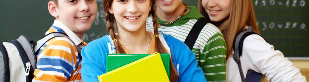 Quatro estudantes lado a lado posando para a foto dentro de sala de aula. Quadro negro ao fundo e os quatro usando mochilas. A menina mais focalizada está abraçando livros