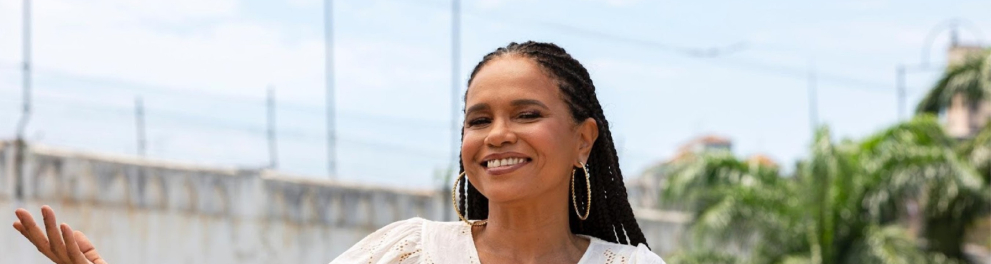 Imagem de pescoço pra cima da cantora brasileira Teresa Cristina à frente de uma paisagem típica do RJ, o bairro de Santa Tereza